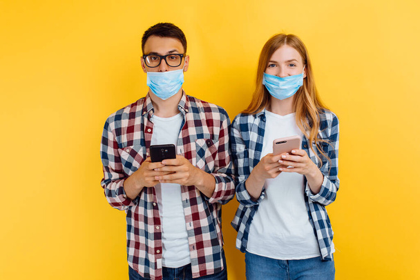 Happy guy and girl in plaid shirts and medical masks on their faces using mobile phones on a yellow background - Foto, immagini