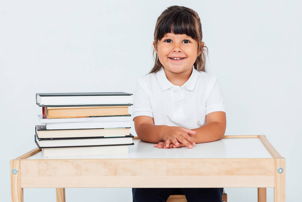 Okuldaki küçük esmer kız, beyaz arka planda kitapları olan bir masanın yanındaki sandalyeye oturmuş gülümsüyor. Okul kavramı - Fotoğraf, Görsel