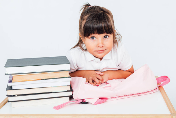 Okuldaki esmer kız, kitaplarla dolu bir masanın yanında oturan, beyaz arka planda pembe sırt çantasına yaslanan. Okul kavramı - Fotoğraf, Görsel
