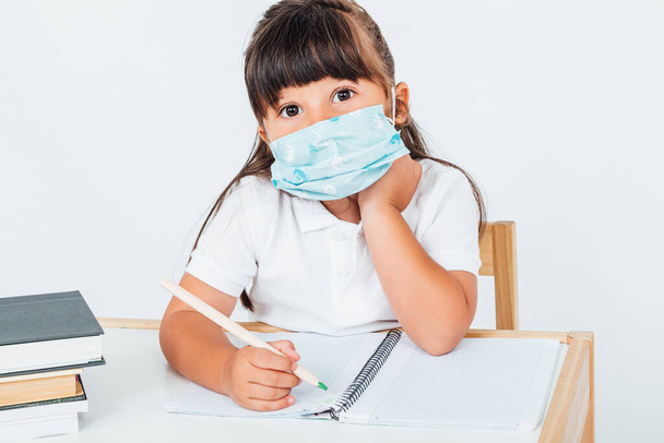 Brunette girl at school wearing a surgical mask to protect herself from COVID-19, sitting in a chair next to a table with books writing, on white background. School concept - Fotoğraf, Görsel