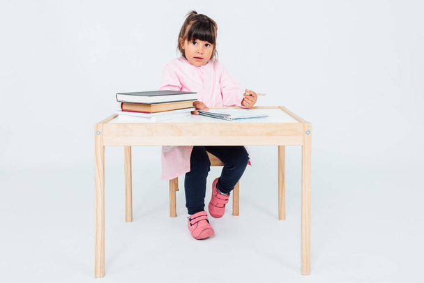 Barna lány az iskolában, komolyan, ül egy széken egy asztal mellett könyvekkel, festéssel, fehér háttérrel. Iskolai koncepció - Fotó, kép