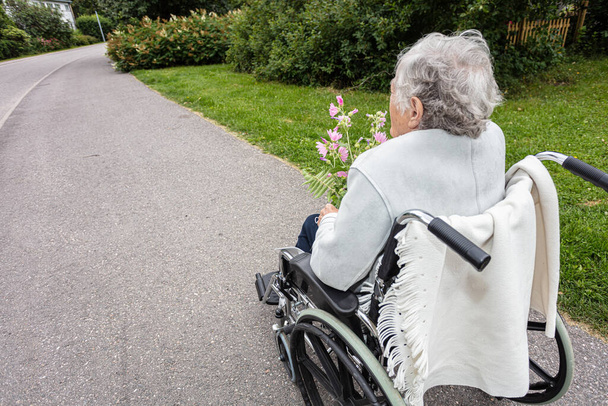 Tekerlekli sandalyedeki yaşlı bir kadın, bahçede çiçeklerle yürürken maske takıyor. - Fotoğraf, Görsel