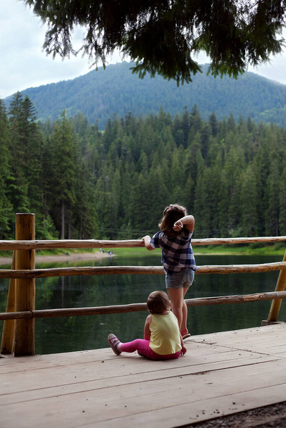 子供たちは美しい山の湖の海岸で遊ぶ。子供は桟橋に座り少女は石を水の中に投げ入れる - 写真・画像