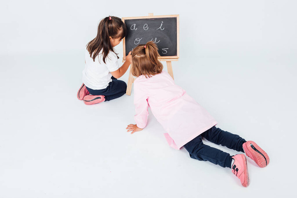 Vista aérea de duas meninas, uma com cabelo preto e outra com cabelo claro, na sala de aula, ao lado de uma tábua de giz, farejando e brincando - Foto, Imagem