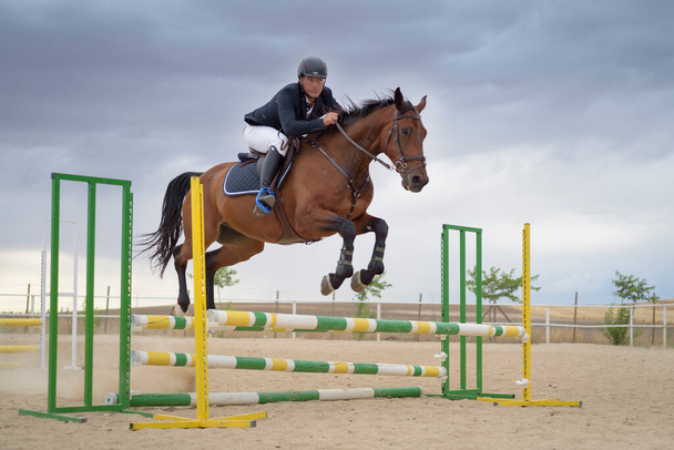 Ο αναβάτης καβαλάει ένα άλογο πηδώντας ένα εμπόδιο στην πίστα. Άλμα αθλητισμού. - Φωτογραφία, εικόνα