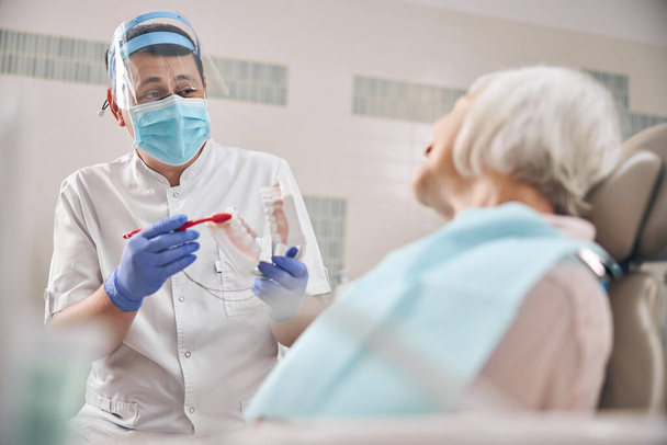 Женщина-дантист разговаривает с пациентом, показывая модель челюсти в стоматологической клинике - Фото, изображение