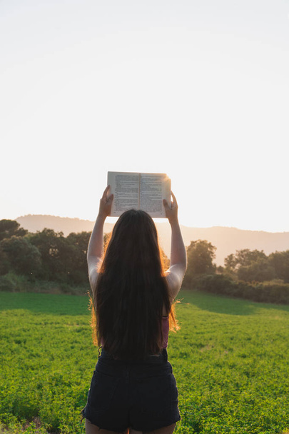 Jong meisje met een open boek in de lucht, op een grasveld bij zonsondergang. Outdoor lezen, onderwijs concept. Vrijheid van geest - Foto, afbeelding
