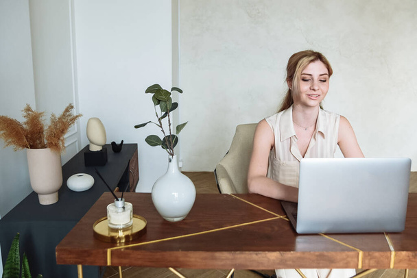 Młoda kobieta za pomocą laptopa, komunikuje się w Internecie z klientem w domu na stole. Przytulne miejsce pracy biurowej, praca zdalna, koncepcja e-learningu. - Zdjęcie, obraz