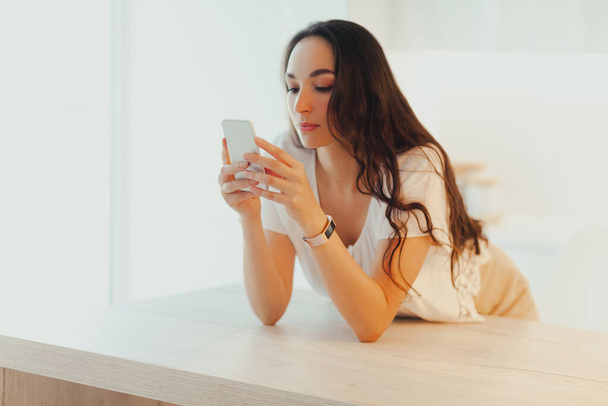 Serious young beautiful woman uses the phone in minimalistic light kitchen. Content creation and communication. Call or message waiting - Foto, Imagem