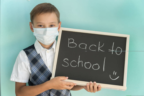 школьник в медицинской маске держит доску с надписью "Вернуться в школу", сделанной из кусочка мела. Копировать место для текста. После года пандемии. Ограничения.  - Фото, изображение