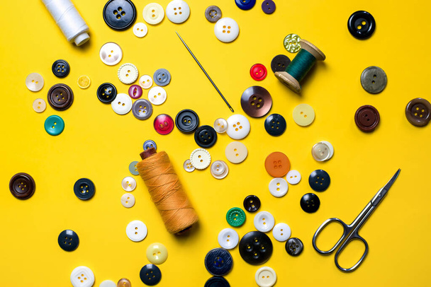 Sewing kit. Multicolored plastic buttons, spools of thread, sewing needles and scissors lie on a bright light yellow background - Zdjęcie, obraz