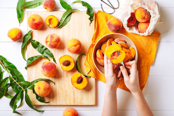 Female hands cutting fresh sweet peaches. Peaches whole fruits leaves, half peach, peach slices on white wooden kitchen table. Recipe making peach jam, cooking peach dessert on cutting board.Flat lay. - Valokuva, kuva