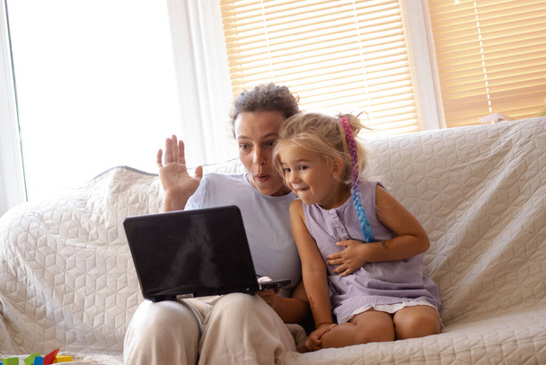 No proszę. Podekscytowana młoda matka z małą uroczą córeczką oglądającą śmieszne kreskówki lub program edukacyjny online na laptopie za pomocą bezprzewodowego łącza internetowego, siedząca razem na kanapie w domu.  - Zdjęcie, obraz