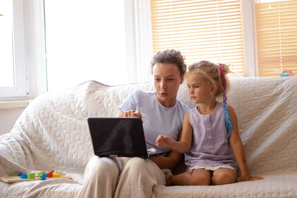 Madre joven enseñando a su hijita cómo usar la computadora portátil, programa educativo en línea, sitio web de navegación, mirando y apuntando a la pantalla del dispositivo electrónico, sentados en el sofá juntos en casa. - Foto, imagen