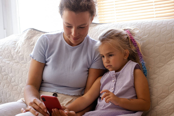 Młoda mama wraz z uroczą córeczką trzymającą smartfona, patrzącą na ekran komórkowy, grającą w aplikację edukacyjną, siedzącą na sofie w domu. Pojęcie kontroli rodzicielskiej. Rodzinny wypoczynek z dzieckiem - Zdjęcie, obraz