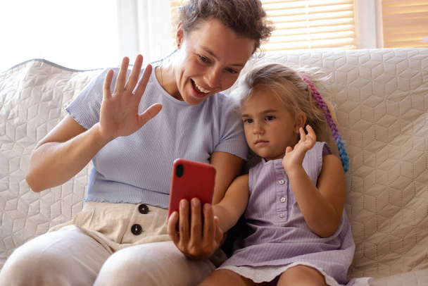 Happy young mother with small daughter making video call with smartphone, gesturing hi, talking with father or grandparents using wireless internet connection, sitting on sofa at home. Communication  - Fotografie, Obrázek