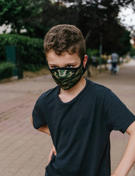 Junge in Militärfarben medizinische Versorgung Maske fahren Roller in einer Fußgängerzone in kurzen Hosen. Neue Normalität. Kindheit. Junge auf einer Walze. Kinder spielen im Freien mit Roller. Aktive Freizeit- und Outdoor-Sportarten für Kinder - Foto, Bild