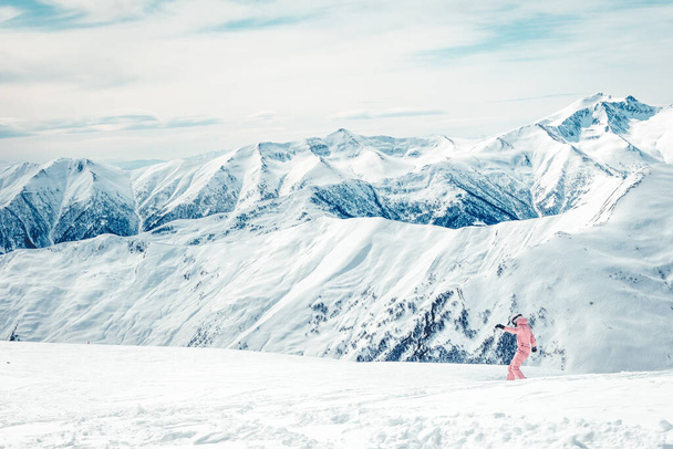 Dziewczyna w różowym kombinezonie zjeżdża ze wzgórza na snowboardzie z widokiem na góry w tle. Urlop narciarski w górach Kazbegi. - Zdjęcie, obraz