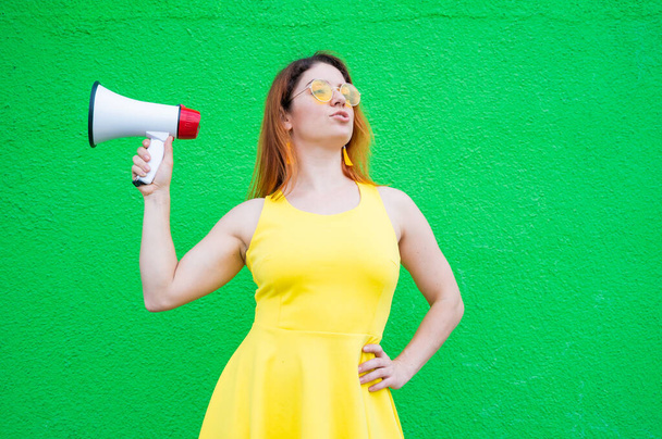 Red-haired woman in sunglasses and a yellow dress holds a megaphone on a green background. - Foto, imagen