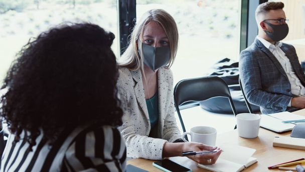 Modern workplace meeting after COVID-19 lockdowns. Young female multiethnic partners talk at office wearing face masks. - Foto, Bild