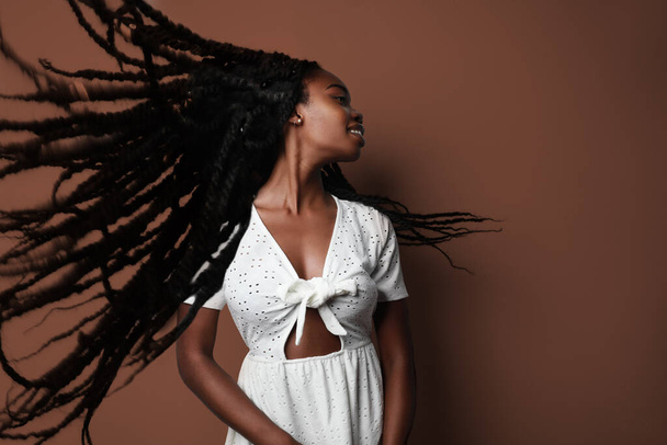 Portrait of young black woman wearing white dress with long hair-locks dancing close to beige background. - Foto, immagini