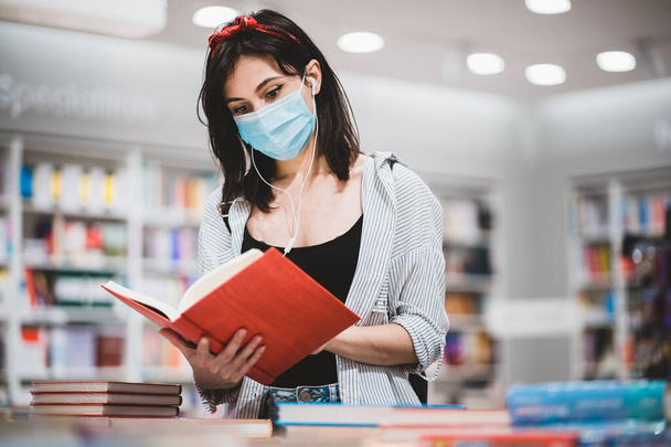 Kafkasyalı öğrenci kız yüz koruma maskesi takıyor kütüphanede kitap okuyor ve kulaklık dinliyor- eğitim, lise, üniversite, öğrenim ve insanlar konsepti - - Fotoğraf, Görsel