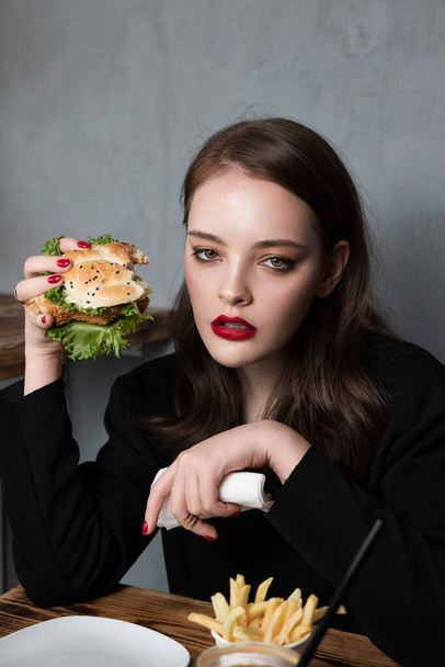 Glamour kobieta z doskonałym moda czerwony manicure i makijaż jedzenia hamburgera i frytki. Koncepcja fast food - Zdjęcie, obraz