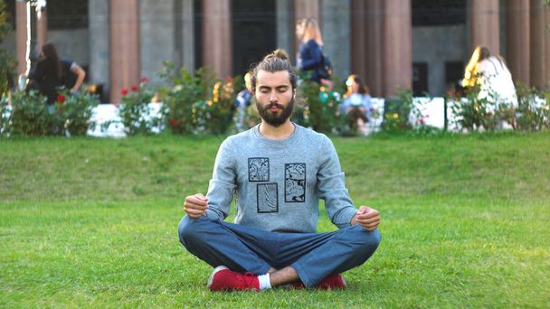 El hombre barbudo medita sobre hierba verde en el parque de la ciudad. Concepto. Elegante joven medita en la hierba verde en la bulliciosa ciudad. Hermoso hombre con estilo está interesado en la meditación - Foto, imagen
