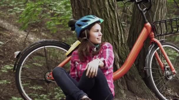 Tema transporte ecológico de bicicletas. Joven mujer caucásica en jeans y estudiante de la camisa se sienta descansando en el parque cerca de la rotura de la bicicleta naranja de alquiler. Chica en la naturaleza descansando después de montar en bicicleta. Descanso después del ciclismo - Imágenes, Vídeo