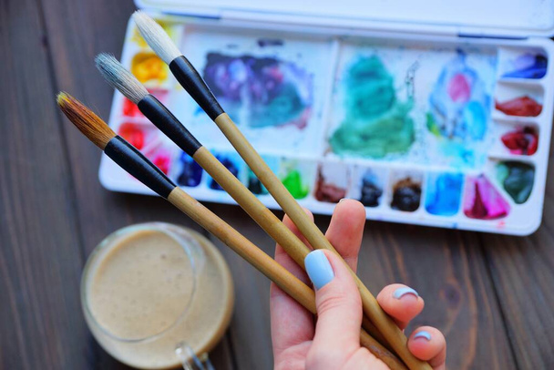 три кисти в руке художника на фоне коробки с цветными красками и стеклянной чашки с коричневым кофе на столе - Фото, изображение
