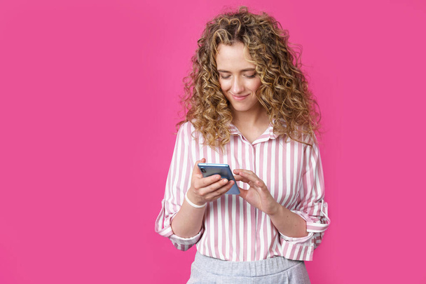 Mujer de moda en una camisa a rayas, escribiendo un mensaje en el teléfono y sonriendo. Aislado sobre fondo rosa - Foto, imagen