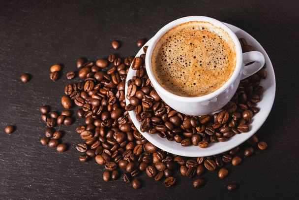 Koffie in een witte beker met een schotel en koffiekorrels op een zwarte achtergrond - Foto, afbeelding