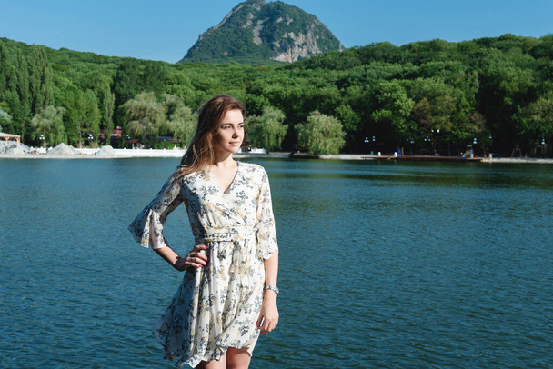 A beautiful and sexy woman, with long hair in a summer dress, spends time on the shore of the lake, surrounded by forests and mountains. Rest and relaxation concept - Valokuva, kuva