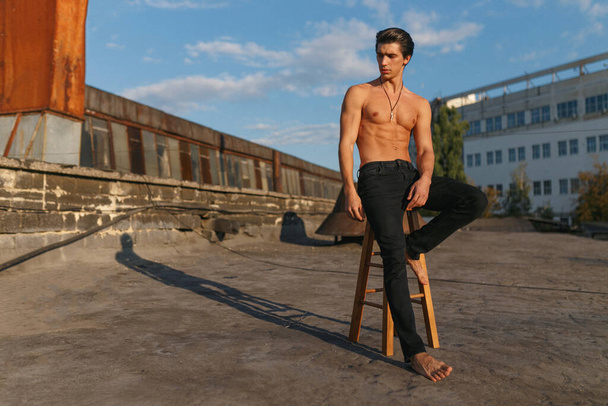 Όμορφος σέξι και αθλητικός νεαρός με γυμνό κορμό, ντυμένος με μαύρο τζιν, ποζάρει σε παλιό εγκαταλελειμμένο εργοστάσιο. Οριζόντια προβολή. - Φωτογραφία, εικόνα