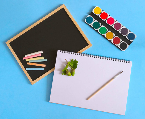 Back to school. Watercolors with white notebook and brush on blue background. Black school chart and chalks. Art concept. - Photo, Image