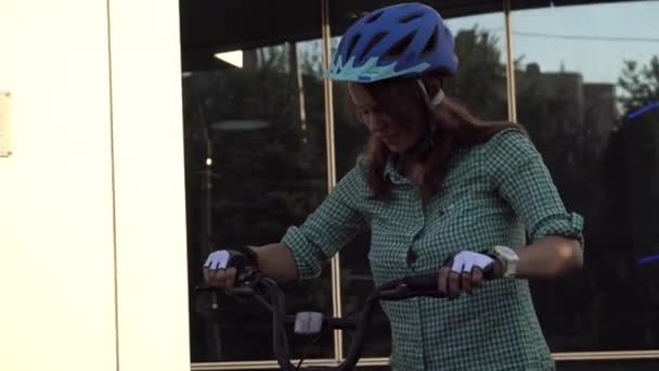 Thème pour travailler sur le vélo. Une jeune femme caucasienne est arrivée au bureau à vélo écologique. Fille dans un immeuble de bureaux de stationnement de vélos dans un casque, gants et chemise et jeans - Séquence, vidéo