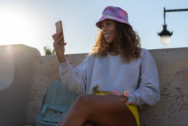 Портрет привлекательной молодой черной женщины, сидящей и разговаривающей с мобильным телефоном на улице без маски. Концепция коммуникации - Фото, изображение
