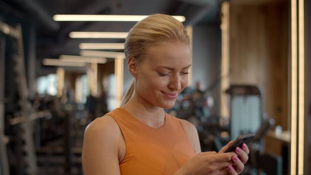 Romantic sportswoman texting message in sport club. Athlete girl standing at gym - 写真・画像