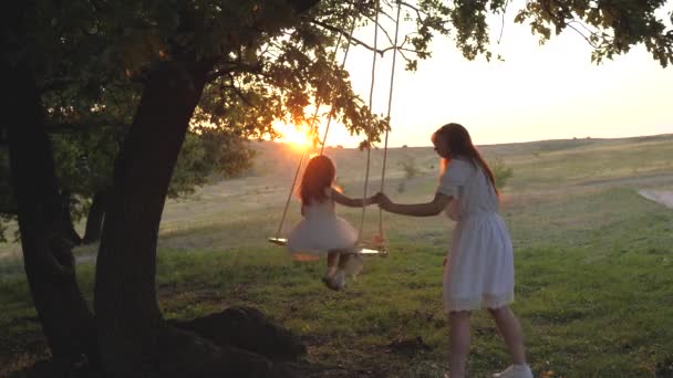 Mutter schüttelt ihre gesunde kleine Tochter auf einer Schaukel unter einem Baum in der Sonne. Mutter spielt mit Kind, sie schwingen am Seil auf einem Eichenzweig im Wald. Die freie Familie hat Spaß im Park. - Filmmaterial, Video