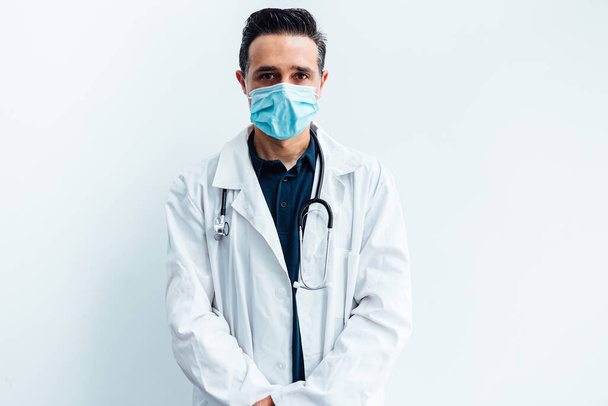 Médico de pelo negro con máscara quirúrgica, con bata blanca y estetoscopio mirando a la cámara, sobre fondo blanco. Concepto de medicina - Foto, Imagen