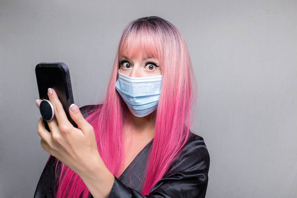 Nainen, jolla on vaaleanpunaiset pitkät hiukset paperinaamarissa, näyttäen yllättyneeltä, lukemalla uutisia sumeasta älypuhelimesta, eristettynä harmaalla taustalla - Valokuva, kuva