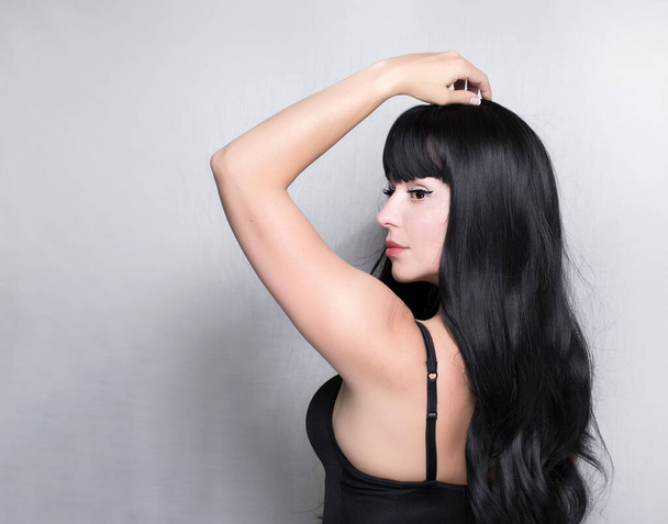 The close up portrait of the caucasian beautiful woman, with black long healthy hair, white skin and red lips, she is holding her hand up on the head, isolated on the grey background - Foto, imagen