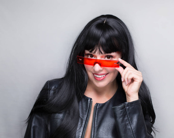 The brunette white woman in the leather jacket, wearing fancy red glasses, smiling, looking forward, isolated on a grey white background - Photo, Image