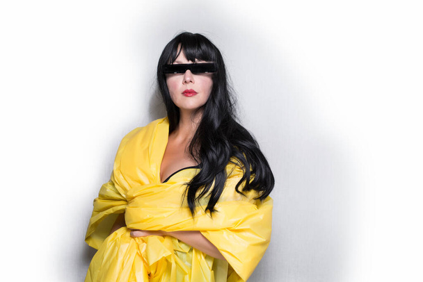 Die brünette weiße Frau in gelber Plastikfolie, mit schicker schwarzer Sonnenbrille, nicht lächelnd, nach vorn blickend, futuristische Mode, isoliert auf grauweißem Hintergrund - Foto, Bild