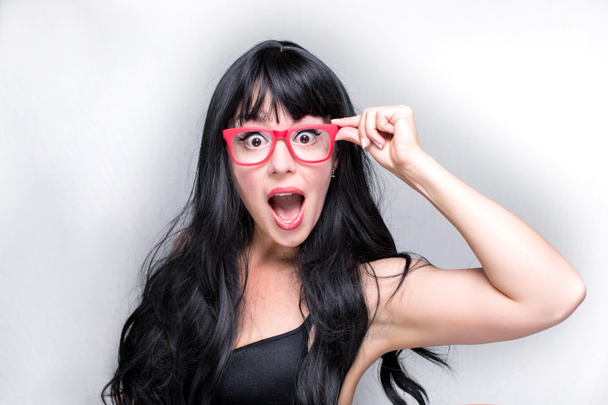 The face of the caucasian young beautiful woman, with black healthy hair, white skin and red eyeglasses, she is touching her eyewear, with open mouth, isolated on the grey background - Fotó, kép