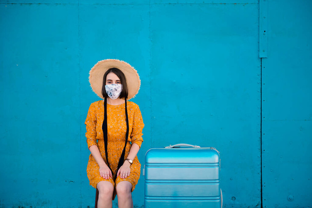 Mujer con un vestido amarillo sentada frente a un fondo azul con su equipaje a su lado - Foto, imagen