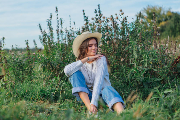 Young beautiful brunette woman dressed in a white sweater, jeans and cowboy straw hat sitting on green grass, smiling and laughing during sunset - Fotó, kép