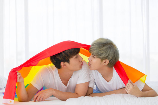 Ασιάτες ομοφυλόφιλοι άνδρες ή γκέι ζευγάρια σε μια ευτυχισμένη στιγμή, ενώ έχουν ένα ουράνιο τόξο σημαία καλύπτει το σώμα και βρίσκεται στο λευκό κρεβάτι. Έννοια της υπερηφάνειας ΛΟΑΤΚΙ. - Φωτογραφία, εικόνα