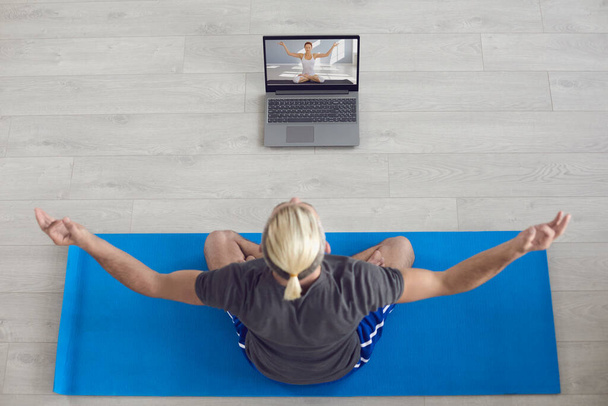 Yoga meditation online. The blond man practices yoga at home. A guy with white hair meditates while sitting on a gray floor in a room. - Fotografie, Obrázek