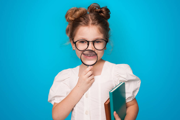 Kis kaukázusi lány szemüveggel, kezében néhány könyvet, és nagyító mosolyog a kék stúdió falán - Fotó, kép